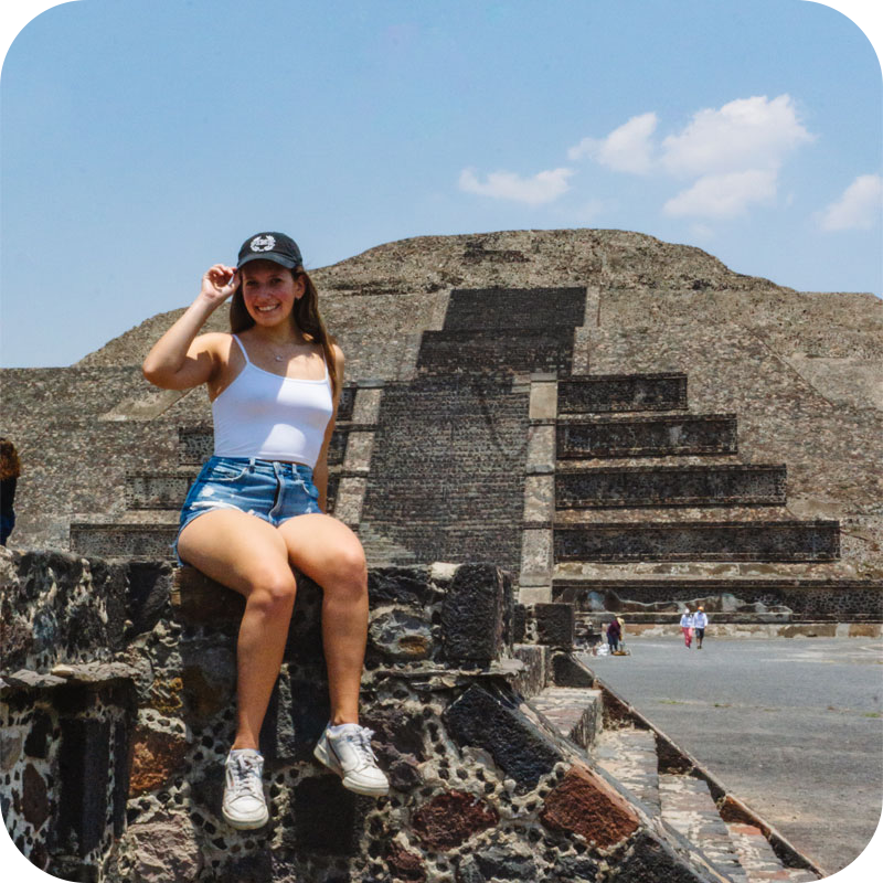 Woman in white shirt and blue denim shorts with hand on hat in front of Teotihuacan ruins in Mexico