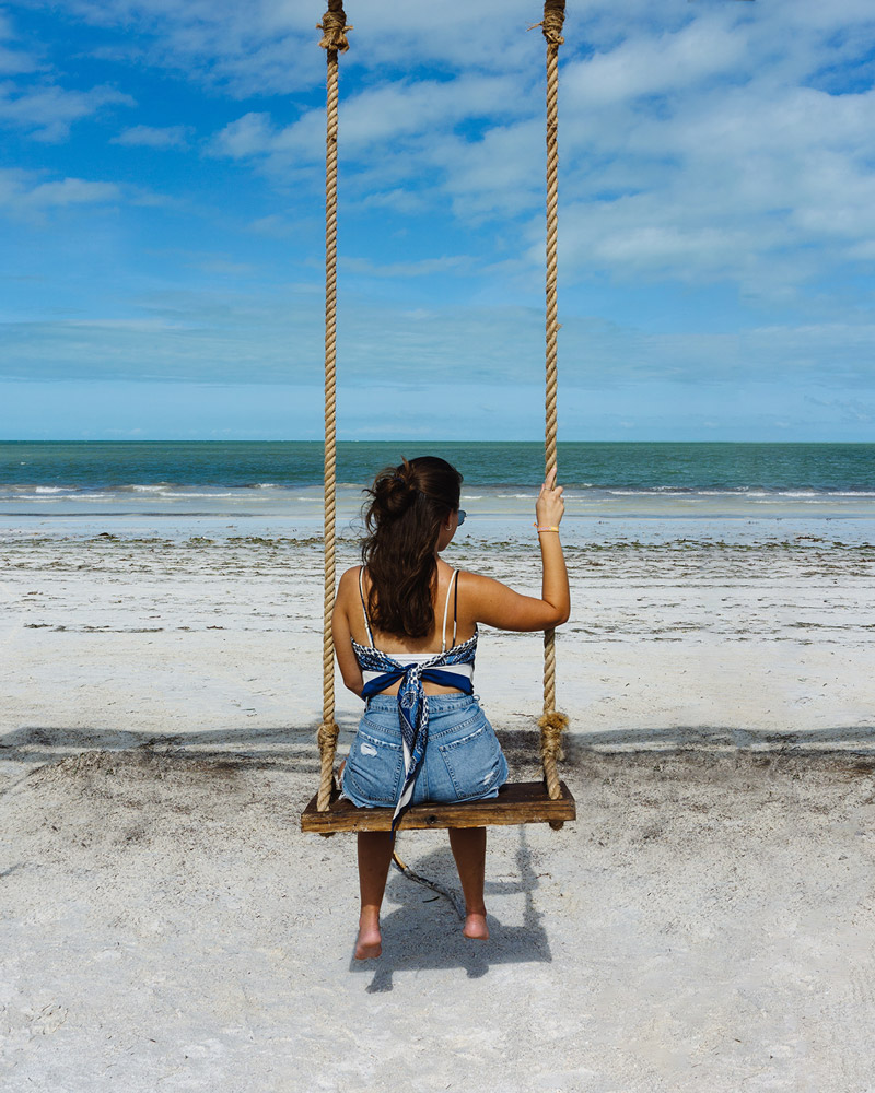 Woman in blue denim shorts and a white and blue shirt on a rope swing on the beach with the ocean and blue sky in the background in Holbox, Mexico