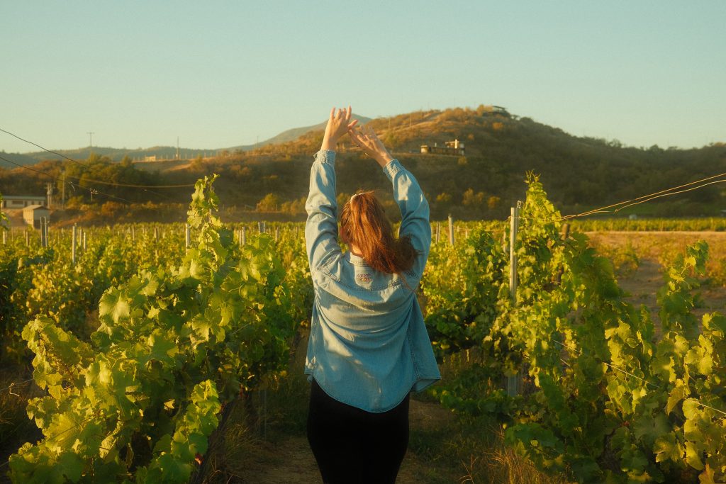 Woman in blue denim shirt and black leggings walking through vineyard in Casablanca, Chile with hands in the air